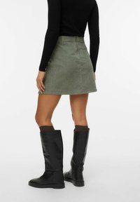 Jupe en velours côtelé kaki avec une forme évasée, assortie à des bottes en cuir noir jusqu'aux genoux et des chaussettes marron foncé, montrant une texture lisse.