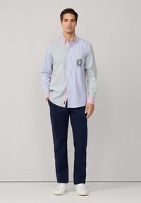 Hombre de pie con las manos en los bolsillos, vistiendo una camisa de botones a rayas multicolores, pantalones azul marino y zapatillas blancas, frente a un fondo liso.