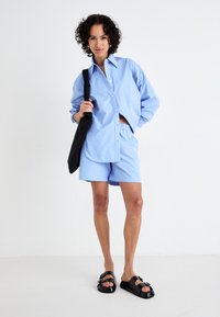 Lys blå skjorte og shorts sett laget av bomull, med en avslappet passform. Svart kviltet veske og svarte slip-on sandaler fullfører looken.