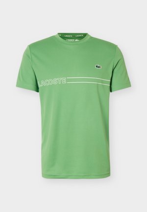 Grøn kortærmet T-shirt med rund hals og teksten "LACOSTE" samt to hvide linjer hen over brystet, med en lille krokodille-logo på højre side.