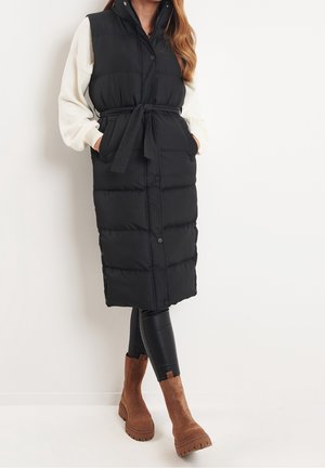 Femme portant un long gilet matelassé noir sans manches avec ceinture à la taille, un haut blanc à manches longues, des leggings noirs et des bottines en daim marron.