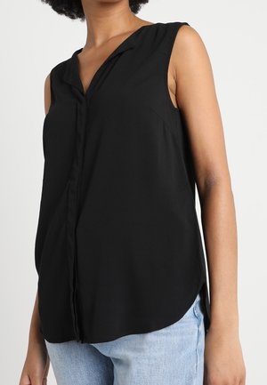 Blouse noire sans manches avec col en V et boutons cachés sur le devant, portée avec un jean bleu clair sur une personne devant un fond uni.