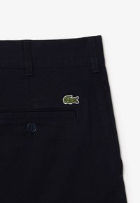 Pantaloni in cotone blu navy con chiusura a bottone e tasca posteriore, caratterizzati da un piccolo logo di coccodrillo ricamato in verde sulla tasca.