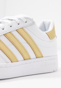 Bílá teniska Adidas se zlatými pruhy, zlatým patníkem, bílými tkaničkami a vyraženým názvem značky na podrážce.