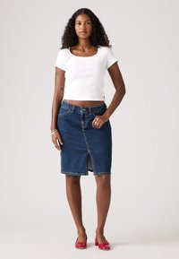 La gonna di denim presenta uno spacco anteriore, chiusura con bottoni e un colore blu indaco. Indossata con un top corto a coste bianco e scarpe rosse.