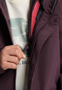 Veste violette avec une fermeture éclair à l'avant et une doublure texturée, portée sur un t-shirt graphique de couleur claire avec des motifs de montagnes et de nuages.