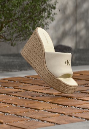 Sandalo con zeppa beige Calvin Klein con piattaforma intrecciata in paglia e punta aperta che fluttua sopra una superficie in legno decorata all'aperto.