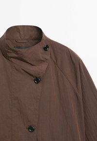 Bruine jas met staande kraag, voorzien van een gestructureerde stof, twee zwarte knopen en een losse pasvorm met schuin lopende naden.