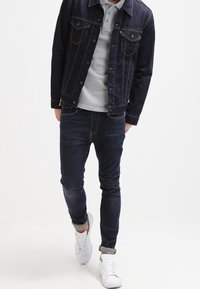 Giacca in denim scuro con cuciture a contrasto, polo grigia, jeans scuri e sneakers bianche. Design semplice, vestibilità casual e maniche arrotolate.