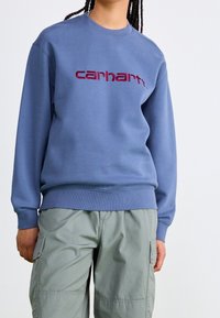 Sudadera azul de algodón con puños y dobladillo acanalados. Presenta un destacado logo bordado "carhartt" en color burdeos en el frente.