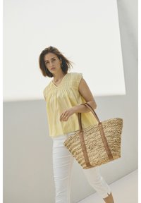 Haut en lin jaune avec une texture plissée, associé à un pantalon blanc et un tote en paille tissée avec des poignées en cuir brun.