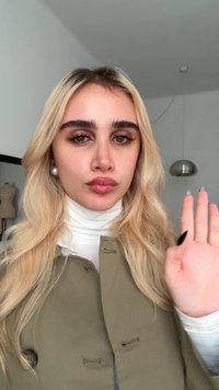 Lichtblond haar, olijfgroene jas met knoopdetails, witte coltrui, hand omhoog houdend, met opvallende wenkbrauwen en glanzende lippen.