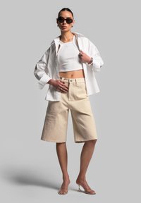 Camicia bianca oversize a bottone sopra un top bianco aderente e pantaloni corti beige da utilità. Gli accessori includono occhiali da sole scuri e tacchi trasparenti.