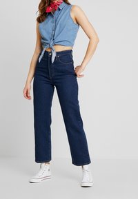 Top en denim court avec un devant noué, associé à un jean bleu foncé taille haute et des baskets blanches. Accent d'un bandana rouge autour du cou.