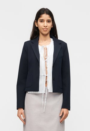 Mujer con blazer azul marino sobre una blusa blanca con lazos y falda gris claro, de pie frente a un fondo blanco liso.