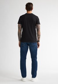 Camiseta de algodón negra, de mangas cortas, cuello redondo, combinada con vaqueros de mezclilla azules, corte recto, y zapatillas blancas, vista desde la parte de atrás.