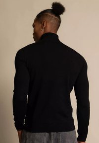 Pull à col roulé noir en tissu côtelé, coupe ajustée, manches longues et col montant avec une texture lisse.