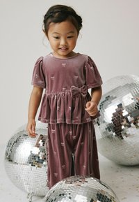 Violette Velour-Zweiteiler mit Puffärmeln, einem Peplum-Oberteil und einer Schleifenapplikation. Die Hose weist ein Schleifenmuster auf. Umgeben von verspiegelten Discokugeln.