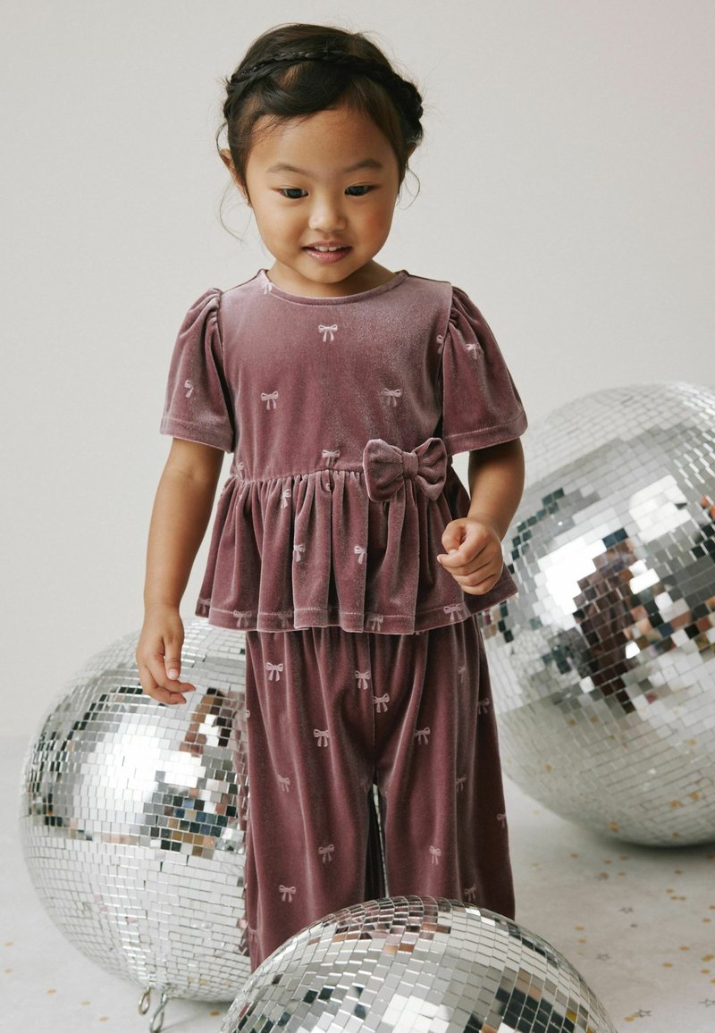 Violette Velour-Zweiteiler mit Puffärmeln, einem Peplum-Oberteil und einer Schleifenapplikation. Die Hose weist ein Schleifenmuster auf. Umgeben von verspiegelten Discokugeln.