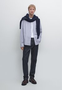 Striped button-up shirt in light blue, worn over a white t-shirt, paired with dark denim trousers and brown lace-up shoes. Navy sweater draped.