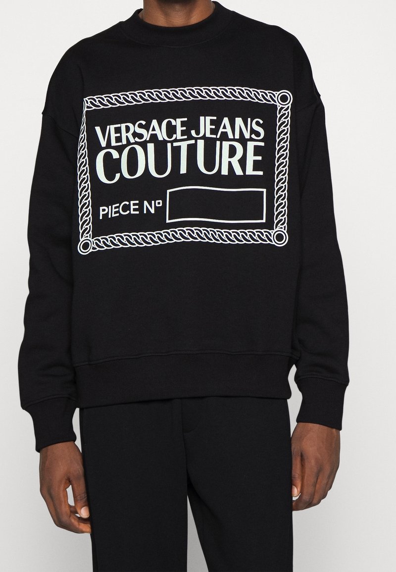 Sweat-shirt noir avec un graphique rectangulaire bordé d'une corde blanche portant l'inscription "Versace Jeans Couture Piece No", porté par une personne à la peau foncée et portant un pantalon noir.