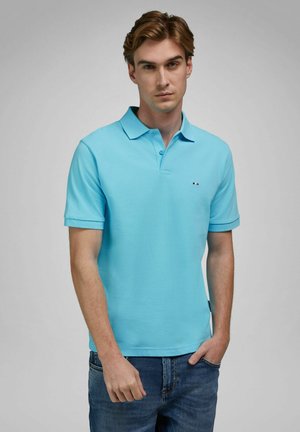 Ung mand med lyst brunt hår iført en lyseblå polo-shirt og blå jeans, stående med den ene hånd i lommen mod en grå baggrund.