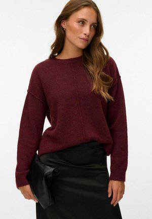 Pull en maille bordeaux à coupe décontractée et manches larges, associé à une jupe en cuir noire ajustée et une pochette noire.