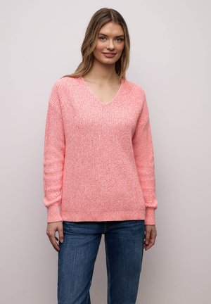 Frau trägt einen hellrosa Strickpullover mit V-Ausschnitt und blaue Jeans, steht vor einem einfarbigen hellen Hintergrund und lächelt leicht.