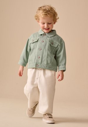 Junges Kind mit lockigem blondem Haar, das eine grüne Jacke, cremefarbene Hose und beige Sneakers trägt, läuft und lächelt vor einem einfarbigen beigen Hintergrund.