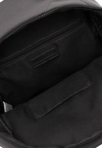 Innenraum einer schwarzen Tasche mit Reißverschlusstasche, eingenähtem Etikett und einer Klappentasche mit sichtbaren Nähten im Hauptfach.