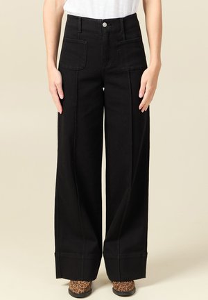 Kobieta ubrana w czarne spodnie o wysokim stanie i szerokich nogawkach z przednimi naszywanymi kieszeniami oraz buty w panterkę, stojąca na beżowym tle.