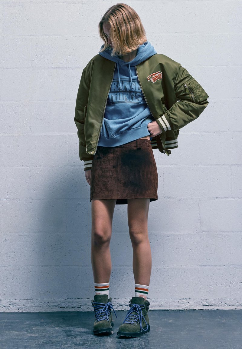 Giubbotto bomber verde oliva con polsini a righe, felpa con cappuccio blu con scritta "Stranger Things", minigonna in camoscio marrone, stivali verdi con lacci.