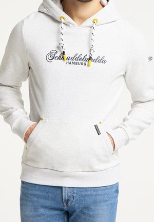 Mand iført lysegrå hættetrøje med sorte og gule snore, "Schruddlewudda Hamburg" tekst og blå jeans, hænderne i forlommerne.