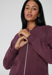 Burgundská mikina na zip s žebrovanými manžetami a kapsami. Má lehkou, texturovanou tkaninu a přiléhavý design límce.