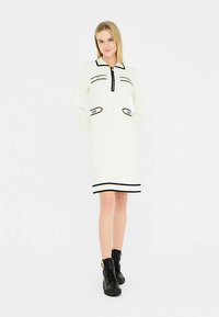 Biele šaty s dlhými rukávmi, čiernym zipsom a detailmi na vreckách; zdobené čiernym lemom na leme a manžetách; skombinované s čiernymi členkovými topánkami.