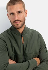 Groene bomberjack met een ritssluiting aan de voorkant, een ribgebreide kraag en een gemêleerde stof. Voorzien van een leren label en een zak op de mouw.