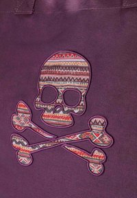 Bolso de tela morado con un gran diseño de calavera y huesos cruzados bordado. La calavera presenta patrones geométricos coloridos y acentos.