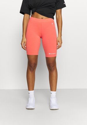 Vrouw in koraalkleurige Champion fietsbroek, zwart cropped T-shirt, witte sokken en witte sneakers, staand tegen een effen lichte achtergrond.