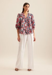 Bluzka w kwiaty w kolorze różowym, granatowym i białym, z dekoltem w kształcie litery V, rękawami o długości trzech kwart oraz luźnym krojem, zestawiona z białymi spodniami o szerokich nogawkach.