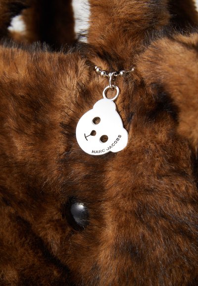 Hnědá taška z umělé kožešiny s kulatou stříbrnou visačkou ve tvaru medvěda se dvěma otvory na oči. Visačka nese nápis "MARC JACOBS."