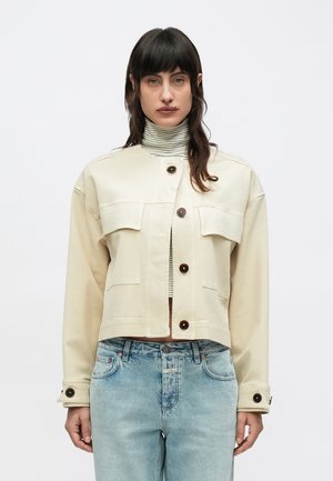 Femme aux cheveux foncés portant une veste courte beige boutonnée, un col roulé rayé et un jean taille haute bleu clair sur fond blanc.