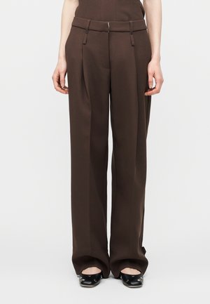 Kobieta nosząca ciemnobrązowe spodnie o szerokich nogawkach i czarne płaskie buty, stojąca na tle jednolitego jasnego tła.