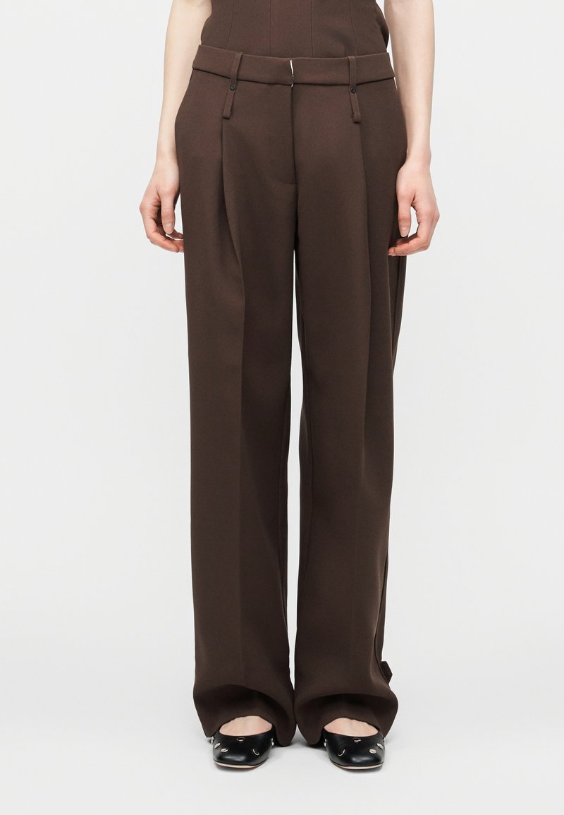 Γυναίκα που φοράει σκούρες καφέ φαρδιές παντελόνες και μαύρα επίπεδα παπούτσια, στέκεται μπροστά σε απλό ανοιχτό φόντο.