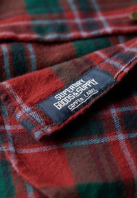 Rood en groen geruit flanellen stof met een zachte textuur, met een blauwe genaaide label waarop "Superdry Goods & Supply Copper Label" staat.