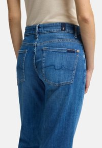 Des jeans en denim bleu présentent une taille haute, deux poches arrières avec des détails de couture et une petite étiquette sur la taille arrière.