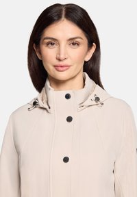 Jasnobeżowa kurtka z wysokim kołnierzem, srebrnymi okuciami i czarnymi guzikami. Charakteryzuje się gładką fakturą i dopasowanym krojem.