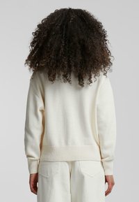 Osoba z kręconymi ciemnymi włosami, ubrana w kremowy sweter i kremowe spodnie, stoi tyłem na jednolitym tle.