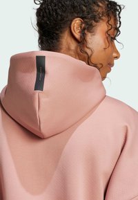 Sudadera rosa con un acabado texturizado, que presenta una gran capucha suave y una etiqueta de logo negra en la parte posterior. Tejido suave con un diseño moderno.