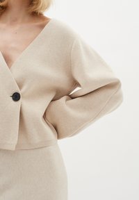 Cardigan bej din tricot cu decolteu în formă de V, mâneci supradimensionate și o închidere cu un singur nasture negru. Textură netedă, croială scurtă și detalii cu tiv ondulat.
