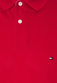 Czerwona koszulka polo wykonana z teksturowanej tkaniny, z kołnierzykiem, dwoma białymi guzikami oraz małym haftowanym logo w kolorze niebieskim, białym i czerwonym.